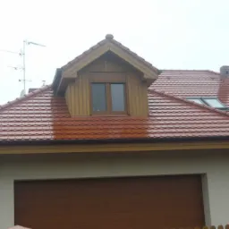 Czerwony dach z lukarną obitą drewnem, widoczne okna dachowe i antena telewizyjna na tle pochmurnego nieba.